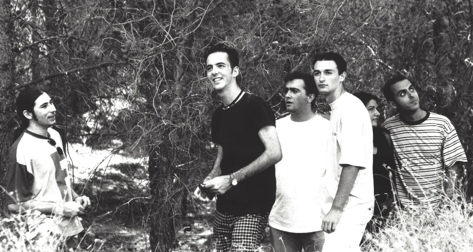 Foto de grupo en blanco y negro de la banda, seis integrantes de pie entre pinos.